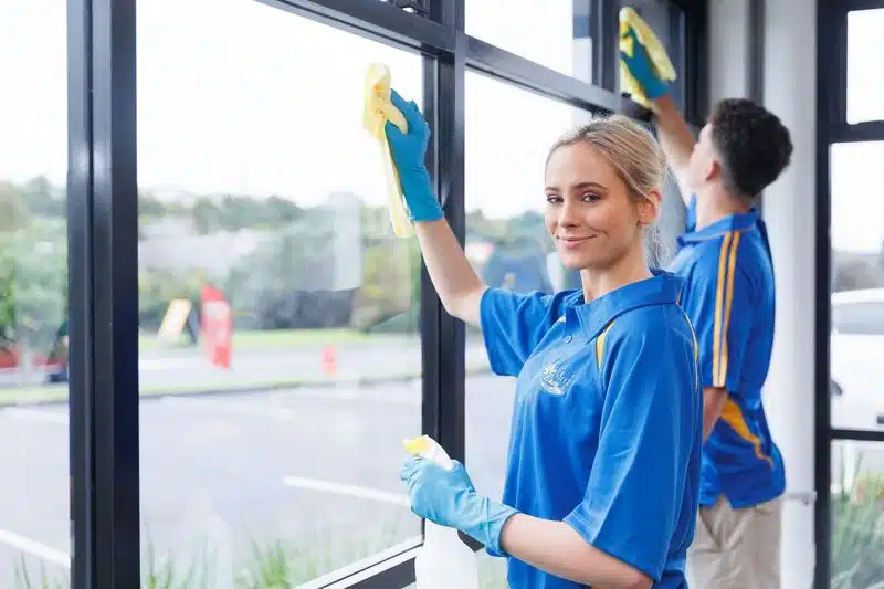 Two people cleaning windows while wearing blue shirts and gloves.