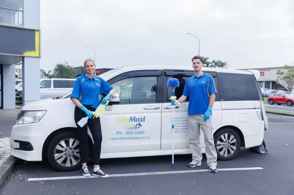 Two people standing beside a white cleaning van, both holding cleaning tools and wearing blue shirts and gloves.