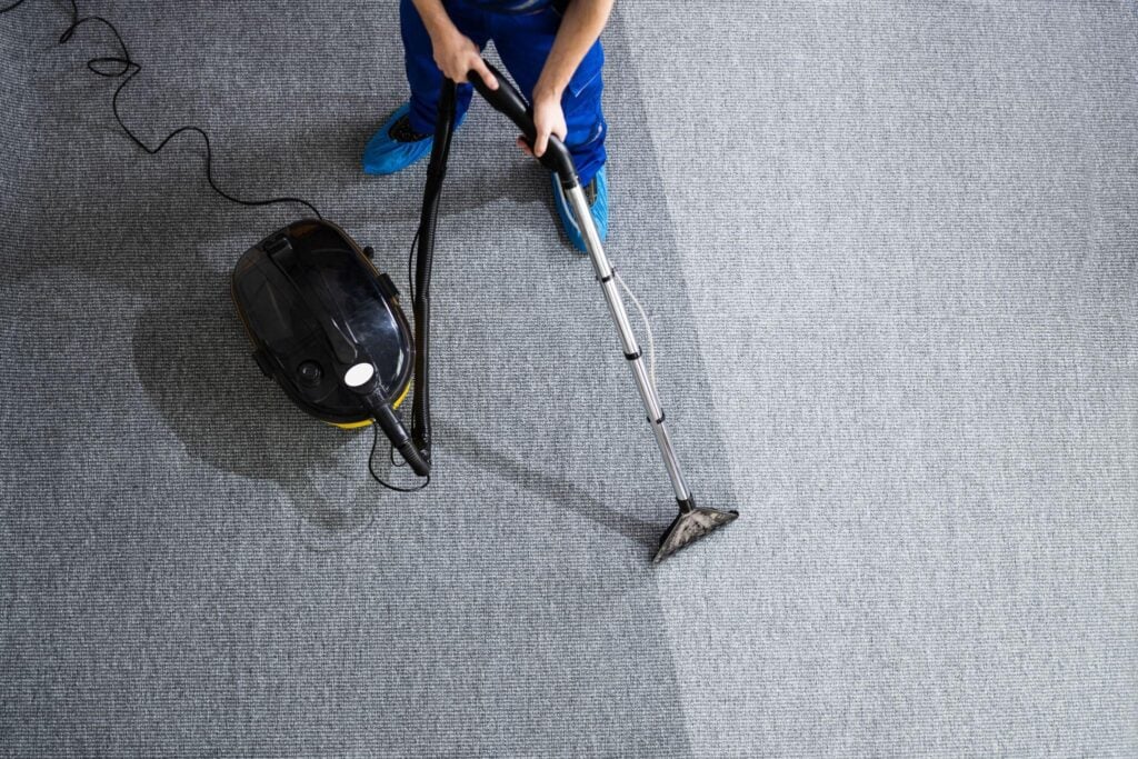 Cleaner using a commercial vacuum machine to remove dust from a carpeted floor.